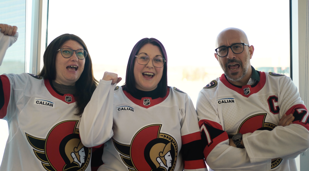 Three people wearing Ottawa Senators hockey jerseys stand indoors, smiling and posing enthusiastically with fists raised.